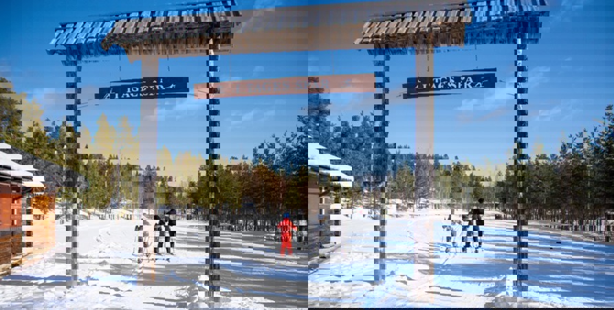 Bilden visar starten vid skidstadion. Tre personer på längdskidor åker under skylten "I Tages spår". 